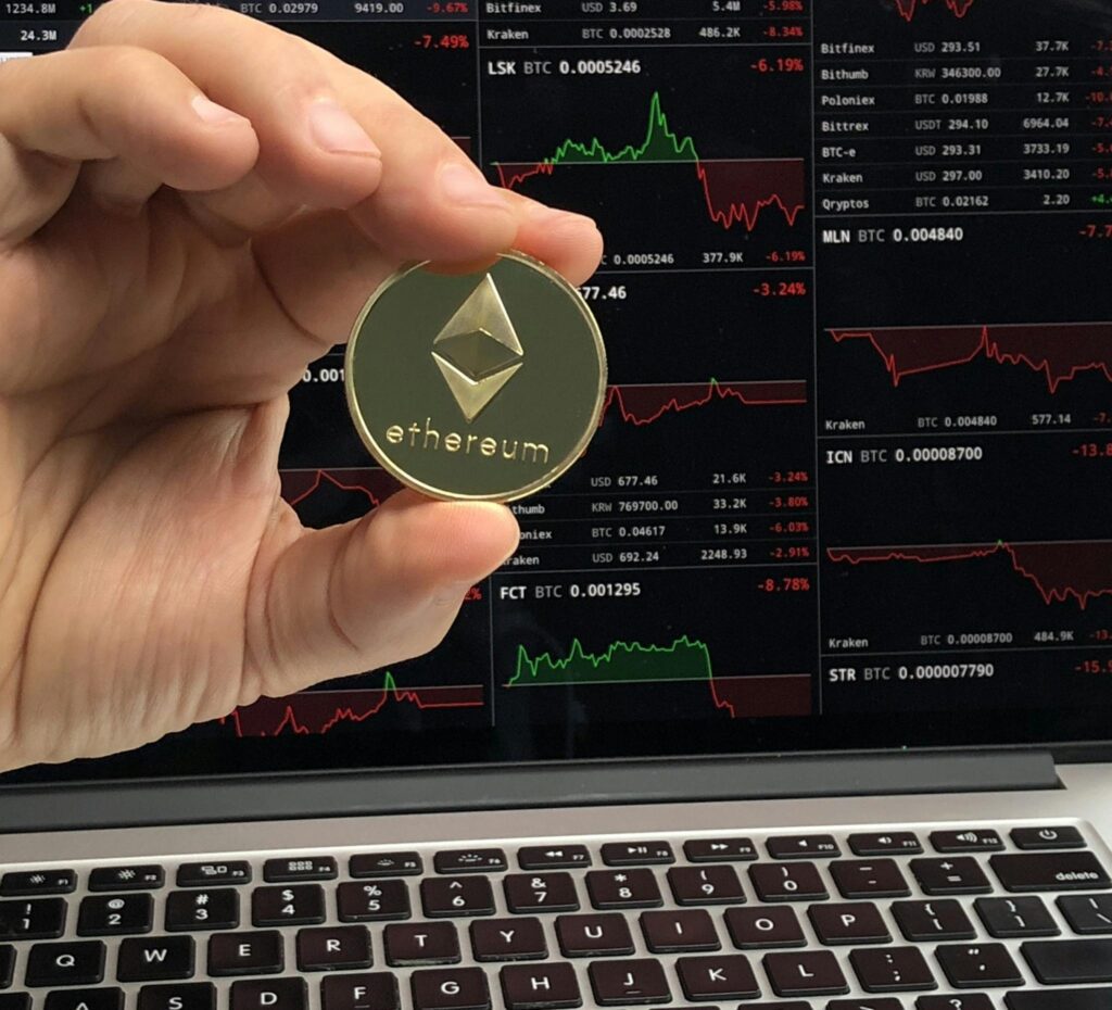 A hand holds an Ethereum coin in front of a laptop displaying cryptocurrency trading charts.